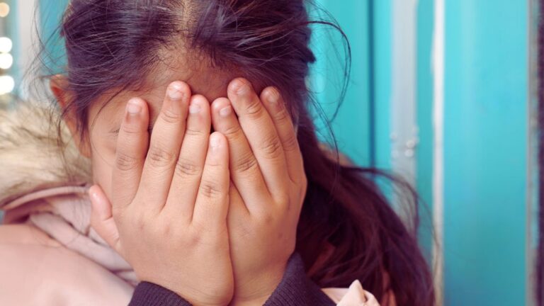 Close-up of a young person with dark hair, wearing a light-colored winter coat with a fluffy fur collar, covering their face with both hands. The background features a bright blue wall and soft bokeh lights, creating a sense of shyness or emotional vulnerability. This image complements the blog 'Autism and Shyness: An ABA Guide,' exploring how ABA therapy supports individuals with autism in managing social anxiety and building confidence.