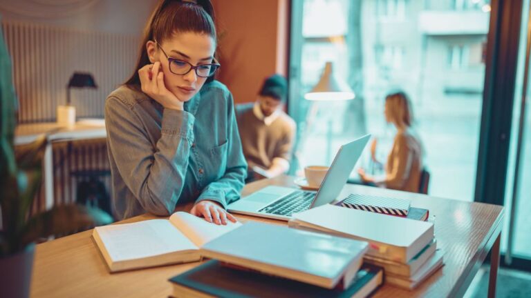 A young woman studying the BCBA exam updates, at a cozy cafe, surrounded by books and a laptop, symbolizes ABA professionals preparing for the BCBA exam updates.