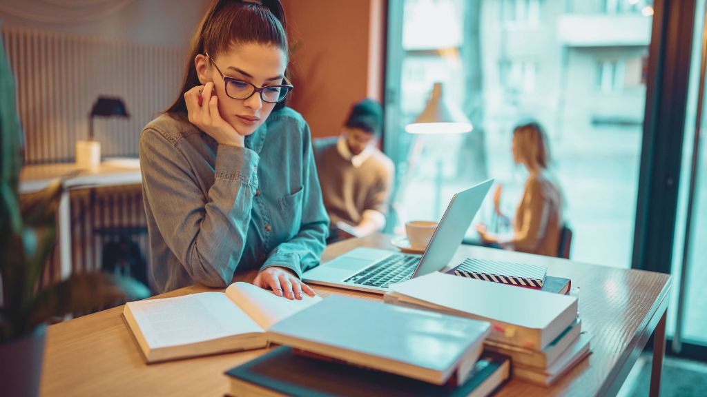 A young woman studying the BCBA exam updates, at a cozy cafe, surrounded by books and a laptop, symbolizes ABA professionals preparing for the BCBA exam updates.