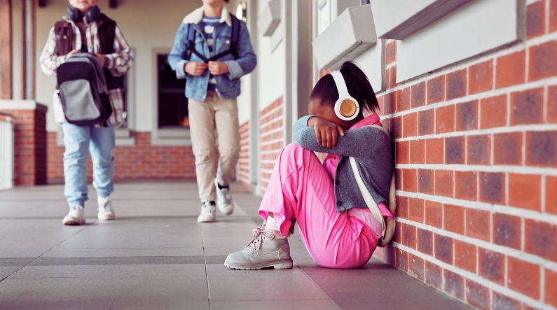 A young girl sits alone against a brick wall, her head buried in her arms, wearing white headphones and pink clothing. Two boys walk away in the background, symbolizing social isolation. This image highlights the challenges of overwhelm and dysregulation in children with autism, as discussed in the blog 'The Window of Tolerance in Children with Autism.'